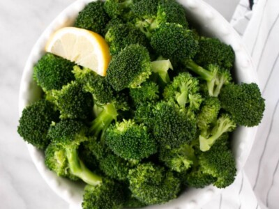 Steamed bright green Broccoli in a white bowl with a slice of fresh lemon.