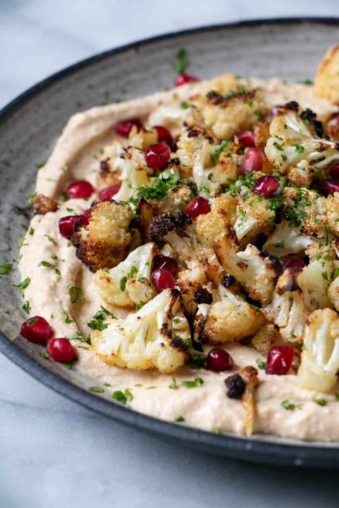 Crispy Cauliflower on a bed of smooth hummus and topped with pomegranate seeds and chopped parsley on a grey ceramic plate - photo taken from an up close side angle. 