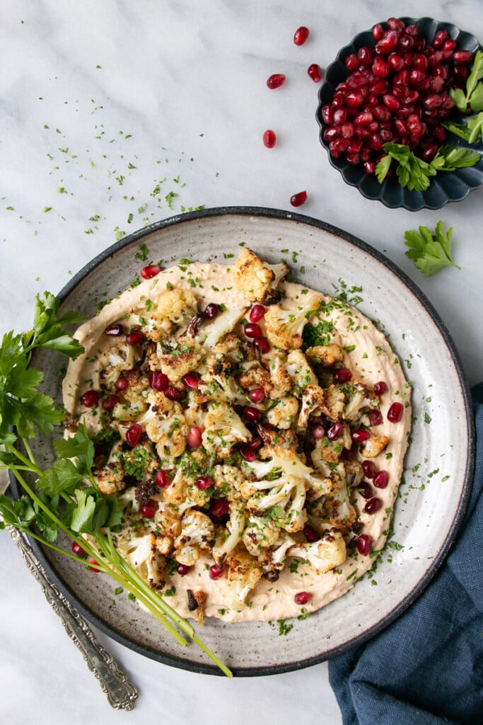 Crispy Cauliflower on a bed of smooth hummus and topped with pomegranate seeds and chopped parsley on a grey ceramic plate and small bunch of parsley stems on the side of the dish and a dark blue side dish pomegranate seeds in it. 