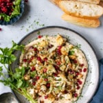 Crispy Cauliflower on a bed of smooth hummus and topped with pomegranate seeds and chopped parsley on a grey ceramic plate and small bunch of parsley stems on the side of the dish and a dark blue side dish pomegranate seeds in it.