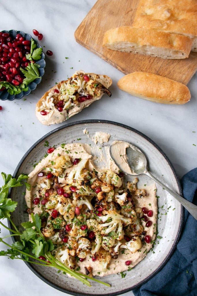 Crispy Cauliflower on a bed of smooth hummus and topped with pomegranate seeds and chopped parsley on a grey ceramic plate and small bunch of parsley stems on the side of the dish with sliced bread topped with the cauliflower hummus dish. 