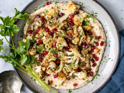 Crispy Cauliflower on a bed of smooth hummus and topped with pomegranate seeds and chopped parsley on a grey ceramic plate and small bunch of parsley stems on the side of the dish.
