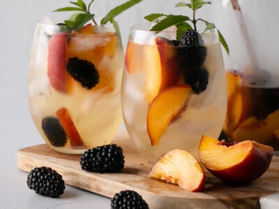 Two stemless wine glasses filled with ice, a white wine based sangria, peaches, blackberries and a bright green fresh mint stem. Blackberries and sliced peaches surround glasses and cutting board.