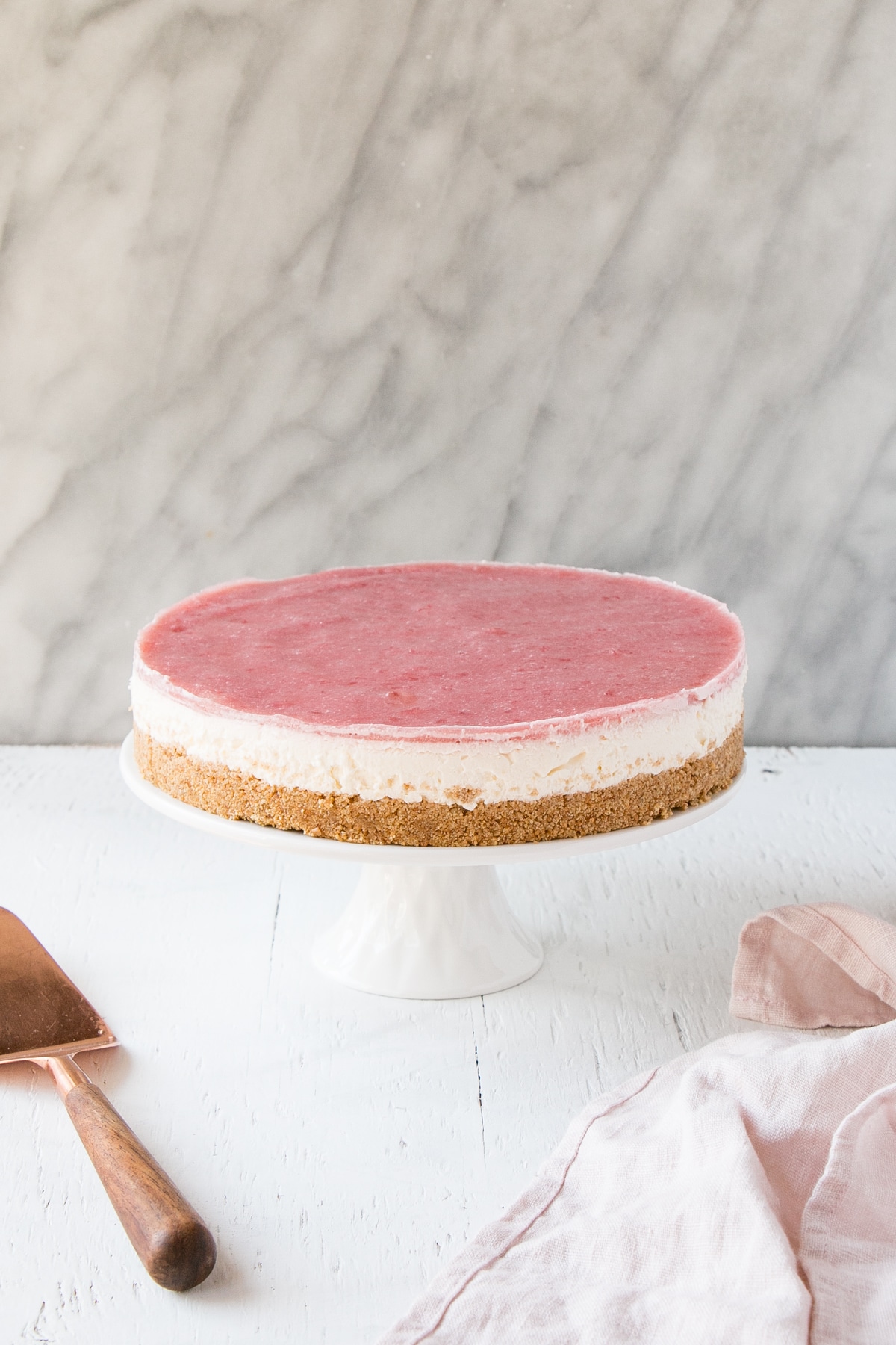 Side view a of No Bake Rhubarb Cheesecake on a white cake stand with a pink tea towel and a serving knife.